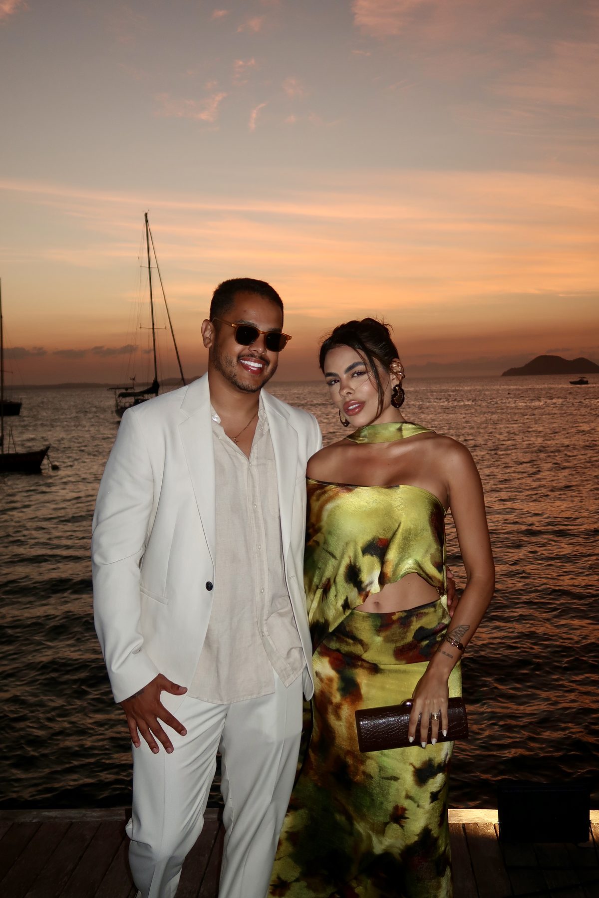 Casal ao pôr do sol em um píer à beira-mar, com barcos ao fundo